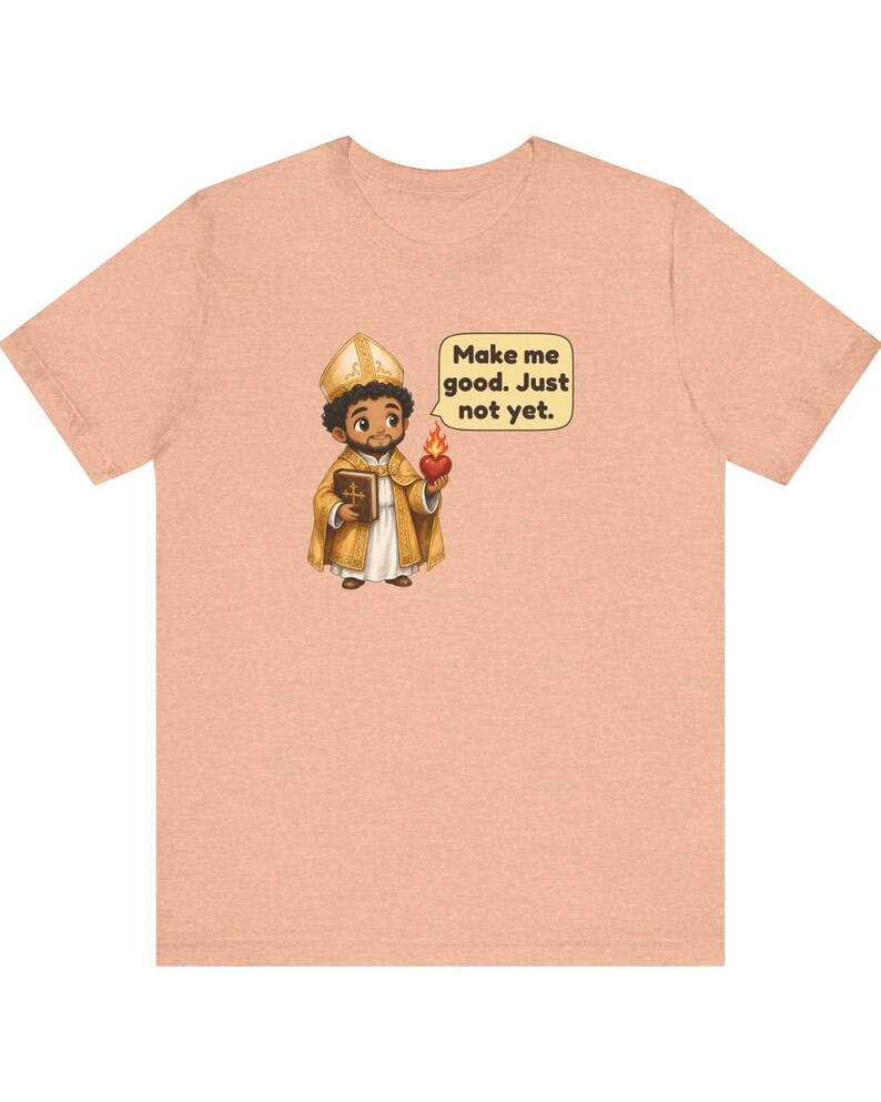 May include: A peach-colored t-shirt featuring a cartoon illustration of a figure in gold religious vestments holding a book and a flaming heart. A speech bubble next to the figure contains the text: Make me good. Just not yet.