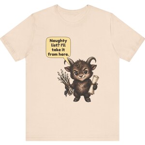 Puede incluir: Una camiseta de color beige claro con el gr&aacute;fico de una peque&ntilde;a criatura marr&oacute;n oscura con cuernos que sostiene un manojo de palos y un pergamino. Un globo de texto amarillo sobre la criatura contiene: Naughty list? I'll take it from here.