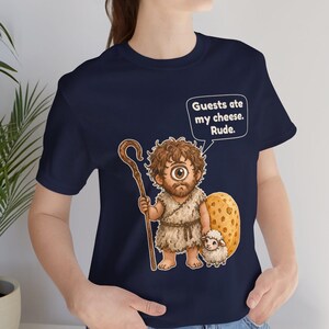 May include: A navy blue t-shirt featuring a graphic of a one-eyed cyclops holding a wooden staff and standing next to a small sheep and a large piece of Swiss cheese. A speech bubble above the character contains the text: Guests ate my cheese. Rude.