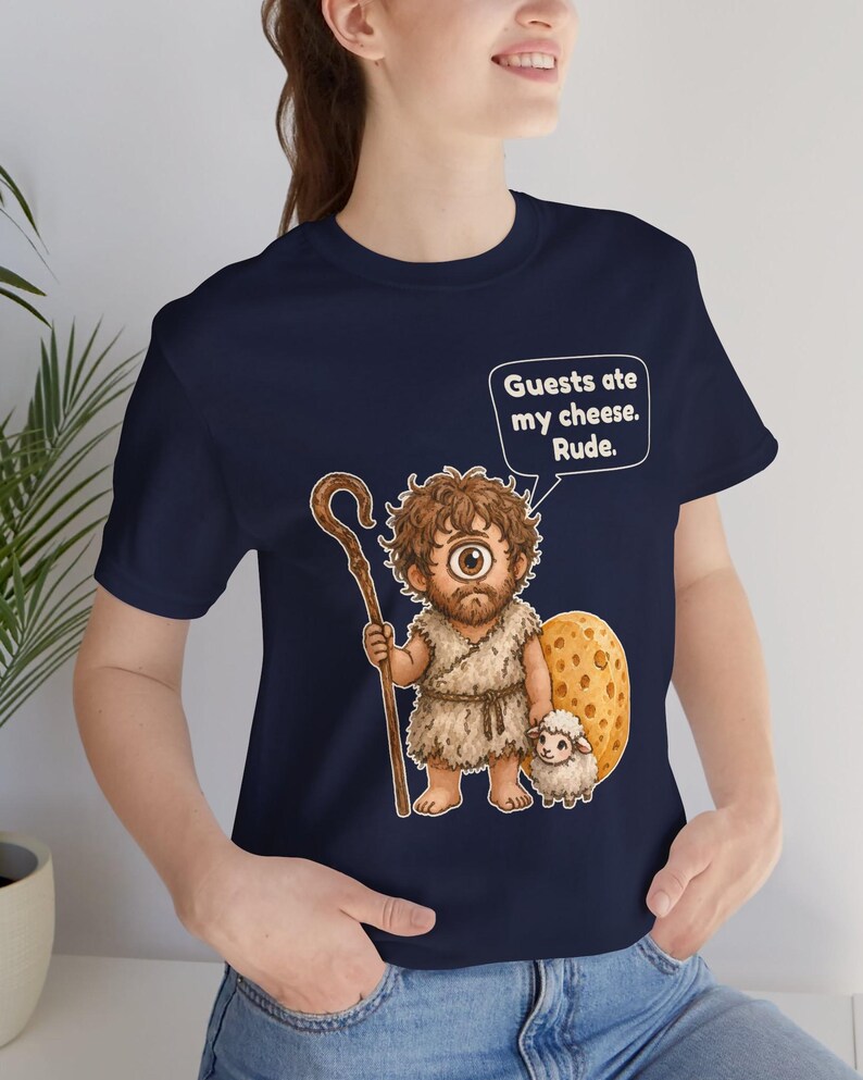 May include: A navy blue t-shirt featuring a graphic of a one-eyed cyclops holding a wooden staff and standing next to a small sheep and a large piece of Swiss cheese. A speech bubble above the character contains the text: Guests ate my cheese. Rude.