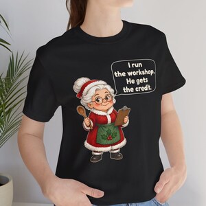 K&ouml;nnte beinhalten: Ein schwarzes T-Shirt mit einer Cartoon-Illustration einer &auml;lteren Frau mit wei&szlig;em Dutt, die ein rotes Kleid mit wei&szlig;em Pelzbesatz, eine gr&uuml;ne Sch&uuml;rze mit Stechpalmen und eine Brille tr&auml;gt. Sie h&auml;lt einen Holzl&ouml;ffel und ein Klemmbrett mit dem Text: I run the workshop. He gets the credit.
