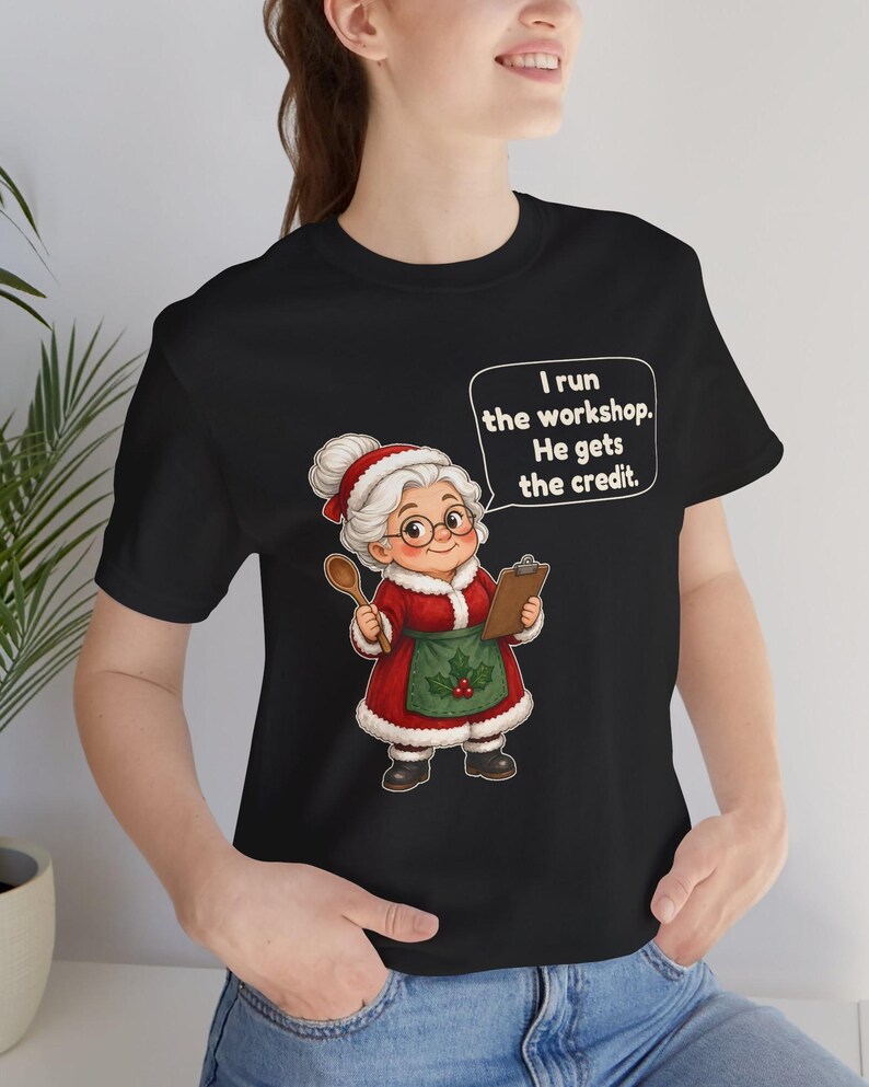 K&ouml;nnte beinhalten: Ein schwarzes T-Shirt mit einer Cartoon-Illustration einer &auml;lteren Frau mit wei&szlig;em Dutt, die ein rotes Kleid mit wei&szlig;em Pelzbesatz, eine gr&uuml;ne Sch&uuml;rze mit Stechpalmen und eine Brille tr&auml;gt. Sie h&auml;lt einen Holzl&ouml;ffel und ein Klemmbrett mit dem Text: I run the workshop. He gets the credit.