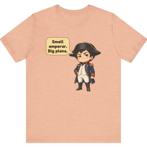 May include: A peach-colored t-shirt featuring a cartoon illustration of a historical figure in a blue military uniform and black hat. A speech bubble next to the character contains the text: Small emperor. Big plans.