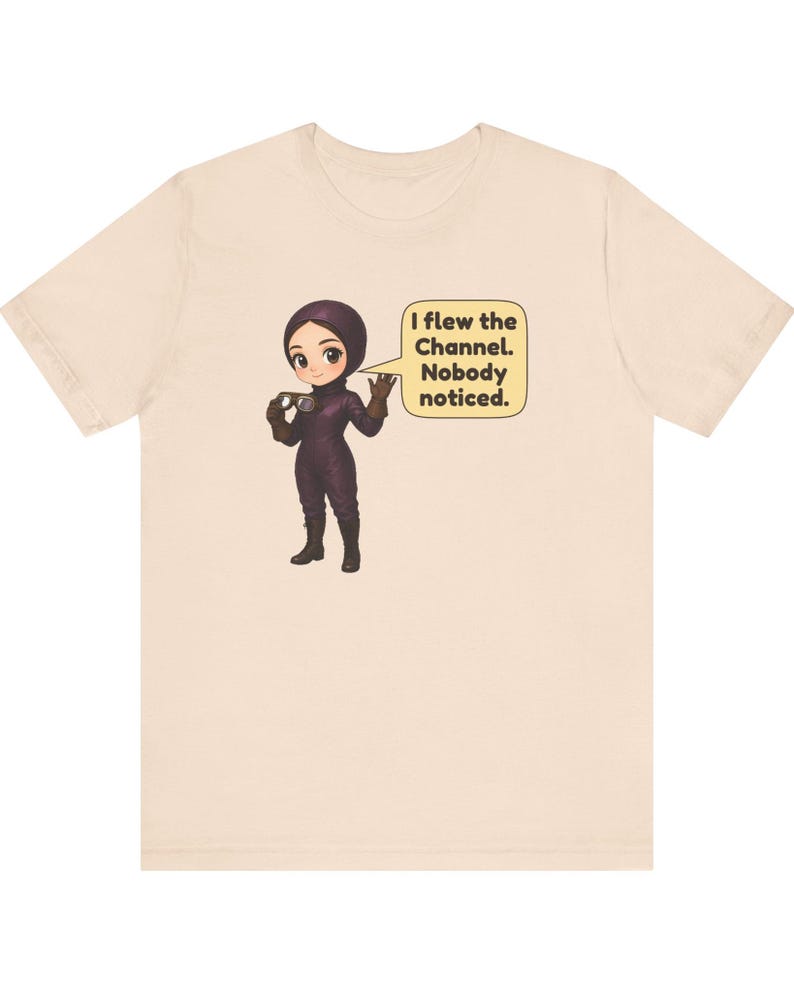 Puede incluir: Una camiseta de color beige claro con una ilustraci&oacute;n de dibujos animados de un personaje con traje de vuelo y casco de color morado oscuro que sostiene unas gafas. Un bocadillo amarillo contiene el texto: I flew the Channel. Nobody noticed.