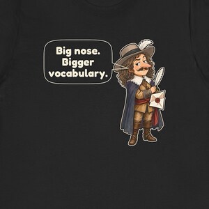 May include: A black t-shirt featuring a cartoon illustration of a historical figure with a large nose, wearing a wide-brimmed hat with a feather, a cape, and boots. The character holds a quill and letter. A speech bubble contains the text: Big nose. Bigger vocabulary.