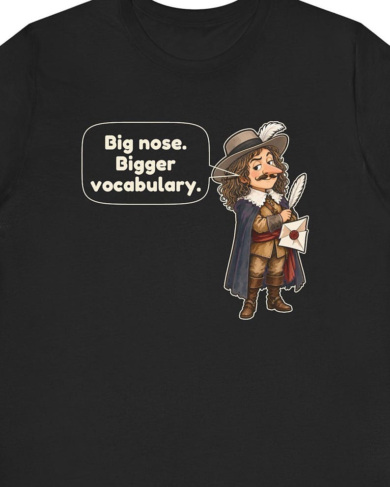 May include: A black t-shirt featuring a cartoon illustration of a historical figure with a large nose, wearing a wide-brimmed hat with a feather, a cape, and boots. The character holds a quill and letter. A speech bubble contains the text: Big nose. Bigger vocabulary.