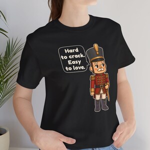 Pu&ograve; includere: Una t-shirt nera a maniche corte con la grafica di un soldatino schiaccianoci in uniforme rossa con dettagli dorati. Accanto al soldato c'&egrave; un fumetto bianco contenente il testo nero: Hard to crack. Easy to love.