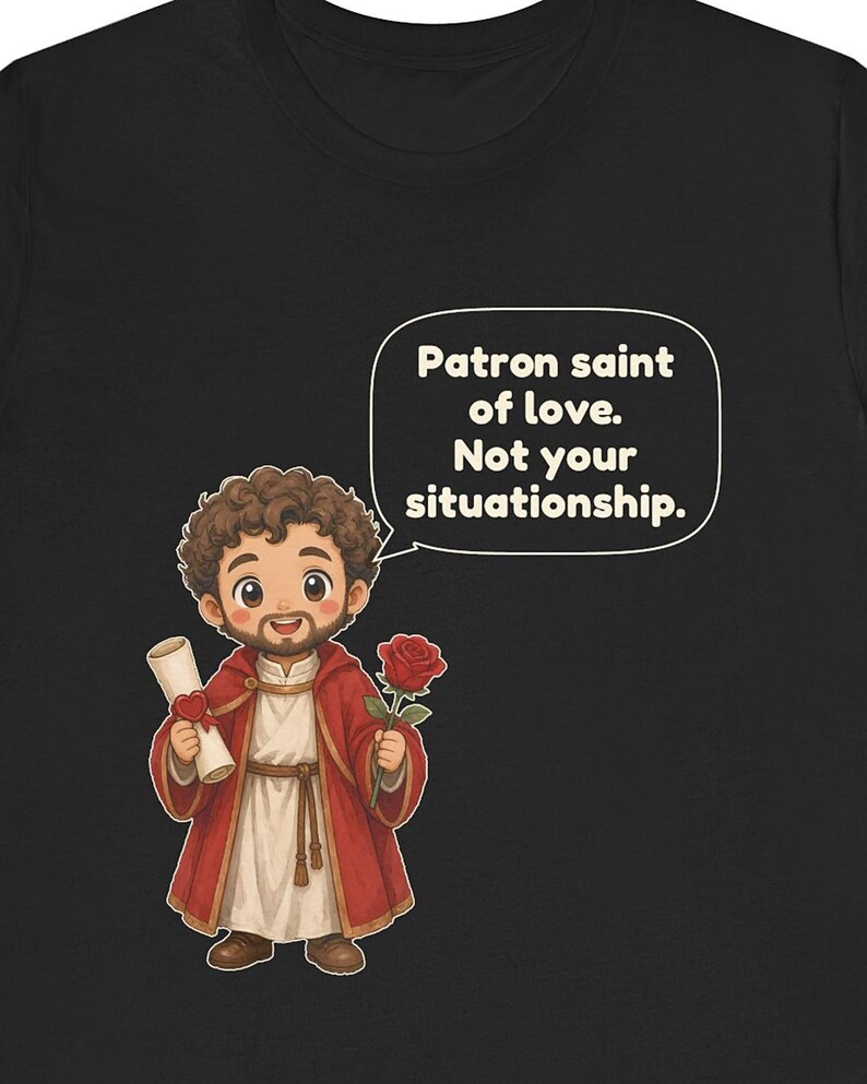 Puede incluir: Una camiseta negra con una ilustraci&oacute;n de dibujos animados de una figura barbuda con t&uacute;nica roja que sostiene un pergamino y una rosa roja. Un bocadillo contiene el texto: Patron saint of love. Not your situationship. El dise&ntilde;o est&aacute; centrado.