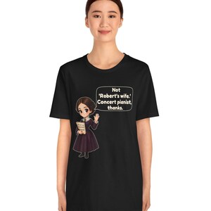 Puede incluir: Una camiseta negra de manga corta con una ilustraci&oacute;n de dibujos animados de una mujer con un vestido morado oscuro sosteniendo un libro. Un globo de texto contiene el texto blanco: Not 'Robert's wife.' Concert pianist, thanks.