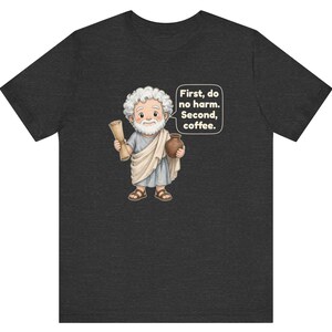 May include: A dark gray t-shirt featuring a cartoon illustration of a bearded figure in a toga holding a scroll and a brown mug. A speech bubble contains the text: First, do no harm. Second, coffee. The design is centered on the front of the shirt.