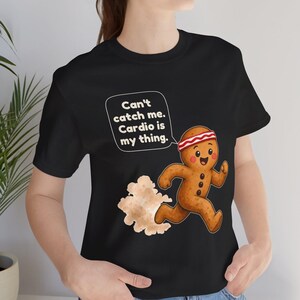 May include: A black t-shirt featuring a graphic of a running gingerbread person wearing a red and white headband. The gingerbread person has a speech bubble that says, "Can't catch me. Cardio is my thing." A cloud of dust trails behind them.