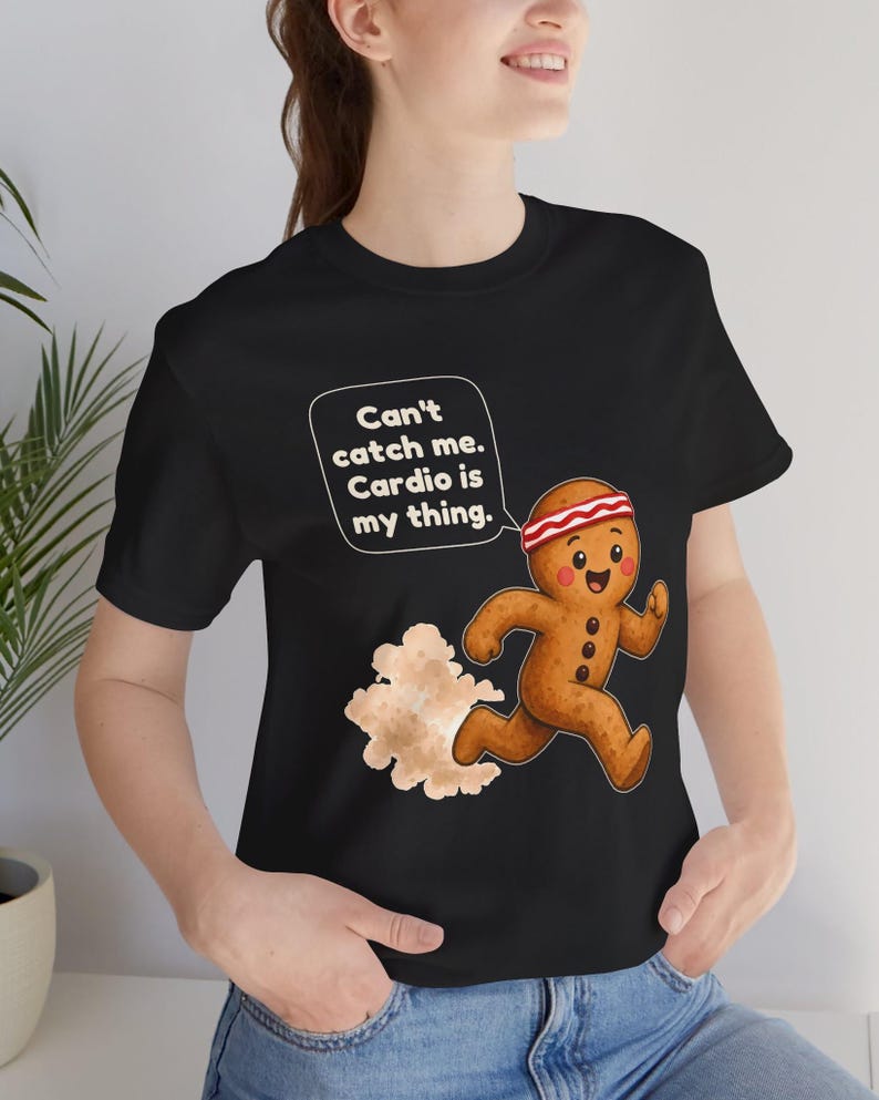 May include: A black t-shirt featuring a graphic of a running gingerbread person wearing a red and white headband. The gingerbread person has a speech bubble that says, "Can't catch me. Cardio is my thing." A cloud of dust trails behind them.