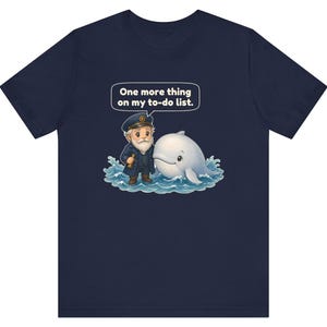 May include: A navy blue t-shirt featuring a graphic of a bearded sea captain in a uniform holding a telescope next to a white whale in blue ocean waves. A speech bubble above them contains the text: One more thing on my to-do list.