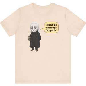 May include: A light beige t-shirt featuring a graphic of a pale, long-haired character in a black coat holding a lit candle. A speech bubble next to the character contains the text: I don't do mornings. Or garlic.