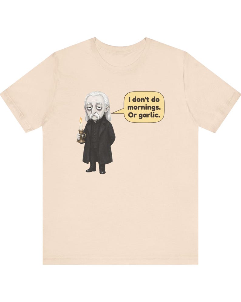 May include: A light beige t-shirt featuring a graphic of a pale, long-haired character in a black coat holding a lit candle. A speech bubble next to the character contains the text: I don't do mornings. Or garlic.