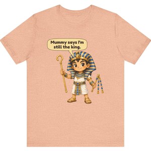 Może przedstawiać: Brzoskwiniowy t-shirt z rysunkową ilustracją młodego faraona trzymającego laskę i bicz. Dymek nad postacią zawiera tekst, Mummy says I'm still the king. Faraon ma na sobie niebiesko-złote nakrycie głowy w paski.