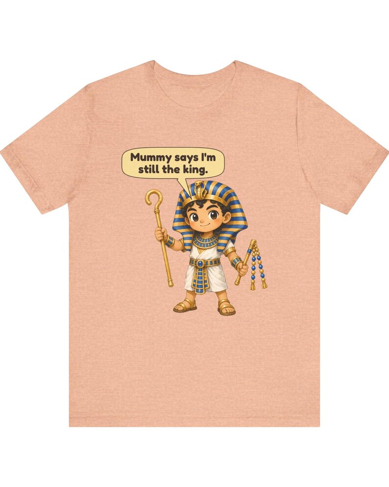 Może przedstawiać: Brzoskwiniowy t-shirt z rysunkową ilustracją młodego faraona trzymającego laskę i bicz. Dymek nad postacią zawiera tekst, Mummy says I'm still the king. Faraon ma na sobie niebiesko-złote nakrycie głowy w paski.