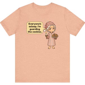Op de afbeelding: Een perzikkleurig t-shirt met een stripfiguur in een rood-wit gestreepte pyjama en bijpassende slaapmuts. Het personage houdt een brandende kaars en een bord met koekjes vast, met een tekstballon: Everyone's asleep. I'm guarding the cookies.