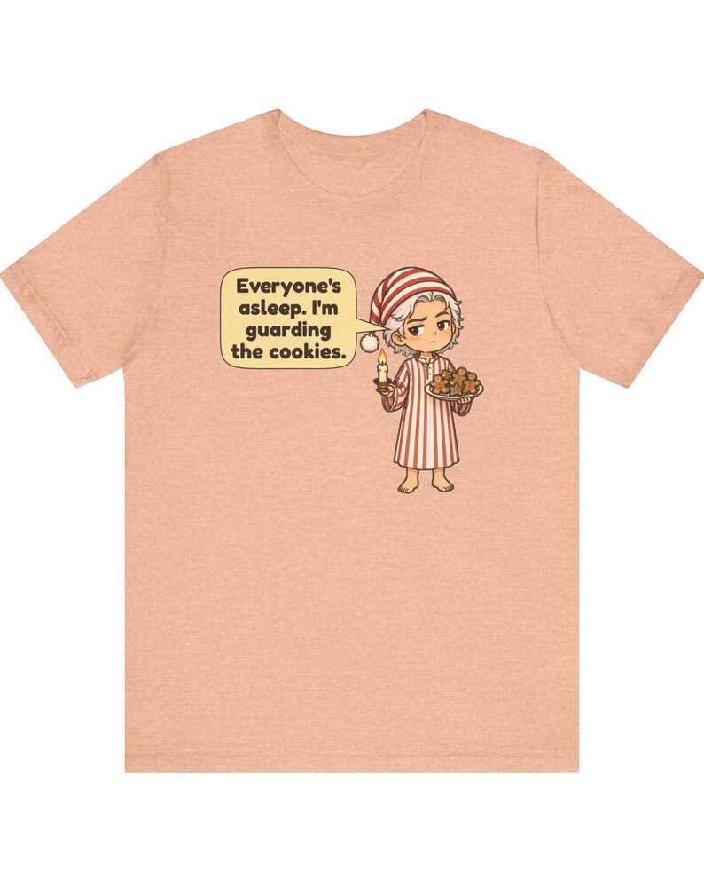 Op de afbeelding: Een perzikkleurig t-shirt met een stripfiguur in een rood-wit gestreepte pyjama en bijpassende slaapmuts. Het personage houdt een brandende kaars en een bord met koekjes vast, met een tekstballon: Everyone's asleep. I'm guarding the cookies.