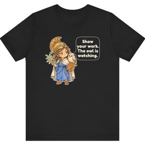 Puede incluir: Una camiseta negra con un gr&aacute;fico de un personaje con casco dorado y t&uacute;nica azul, sosteniendo una rama de olivo y un portapapeles con un b&uacute;ho en el hombro. Un globo de texto contiene: Show your work. The owl is watching.