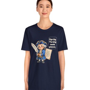 May include: A navy blue short-sleeve t-shirt featuring a cartoon illustration of a historical figure in a blue coat and tricorn hat holding a quill and parchment. A speech bubble contains the text: Sign big. The king needs glasses.
