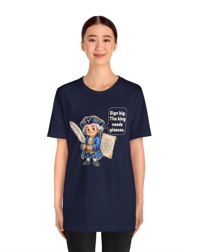 May include: A navy blue short-sleeve t-shirt featuring a cartoon illustration of a historical figure in a blue coat and tricorn hat holding a quill and parchment. A speech bubble contains the text: Sign big. The king needs glasses.
