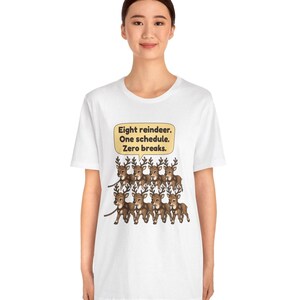 K&ouml;nnte beinhalten: Ein wei&szlig;es Kurzarm-T-Shirt mit einer Grafik von acht braunen Rentieren in zwei Reihen. &Uuml;ber den Rentieren befindet sich eine gelbe Sprechblase mit dem Text: Eight reindeer. One schedule. Zero breaks.