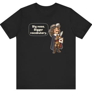 May include: A black short-sleeved t-shirt featuring a cartoon illustration of a historical figure with a large nose, wearing a hat and cape, holding a quill and letter. A speech bubble contains the text: Big nose. Bigger vocabulary.