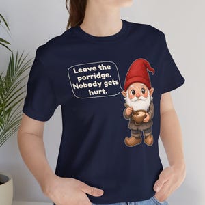 May include: A navy blue t-shirt featuring a graphic of a gnome wearing a red hat and brown clothing, holding a bowl of porridge with a spoon. A speech bubble next to the gnome contains the text: Leave the porridge. Nobody gets hurt.