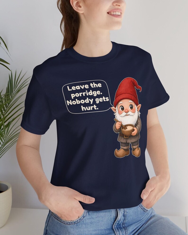 May include: A navy blue t-shirt featuring a graphic of a gnome wearing a red hat and brown clothing, holding a bowl of porridge with a spoon. A speech bubble next to the gnome contains the text: Leave the porridge. Nobody gets hurt.