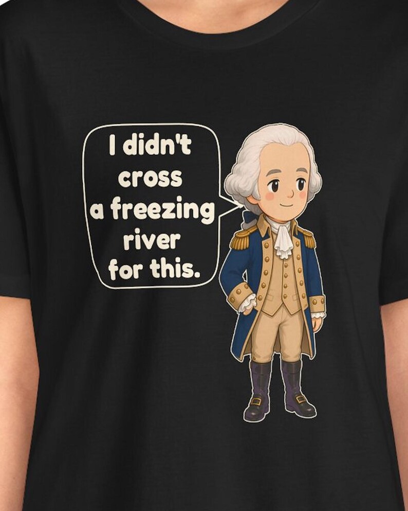 May include: A black t-shirt featuring a cartoon illustration of a historical figure in a blue coat, tan vest, and white cravat. A speech bubble next to the figure contains the text: I didn't cross a freezing river for this.