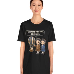 May include: A black t-shirt featuring a graphic of two historical figures in 18th-century attire standing next to a hot air balloon with a sheep inside. A speech bubble above them contains the text: The sheep flew first. Obviously.