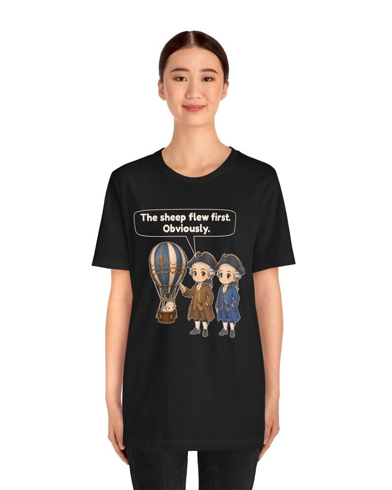 May include: A black t-shirt featuring a graphic of two historical figures in 18th-century attire standing next to a hot air balloon with a sheep inside. A speech bubble above them contains the text: The sheep flew first. Obviously.