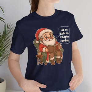 Op de afbeelding: Een marineblauw t-shirt met een afbeelding van de Kerstman die in een groene fauteuil zit en een boek leest, met een mok op een bijzettafel. Een tekstballon naast de Kerstman bevat de tekst: Ho ho hold on. Chapter ending.