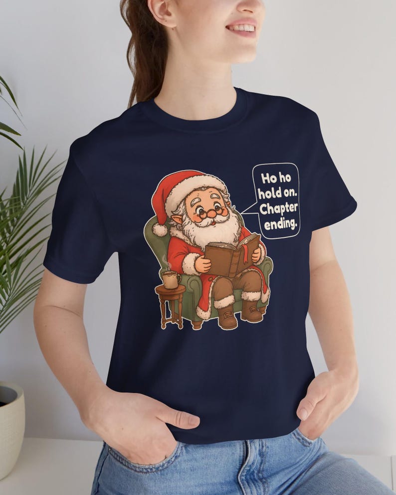 Op de afbeelding: Een marineblauw t-shirt met een afbeelding van de Kerstman die in een groene fauteuil zit en een boek leest, met een mok op een bijzettafel. Een tekstballon naast de Kerstman bevat de tekst: Ho ho hold on. Chapter ending.