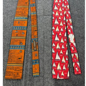 May include: Two neckties displayed side by side. The left tie is orange with blue horizontal stripes, featuring nautical symbols and text about international Morse code. The right tie is red with a repeating pattern of white sailboats and text.