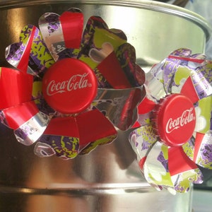 Puede incluir: Dos flores de tap&oacute;n de botella de Coca-Cola rojas y plateadas con detalles verdes y morados.