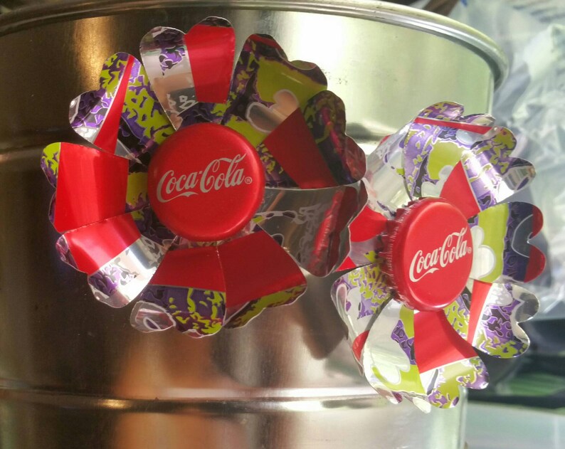 Puede incluir: Dos flores de tap&oacute;n de botella de Coca-Cola rojas y plateadas con detalles verdes y morados.