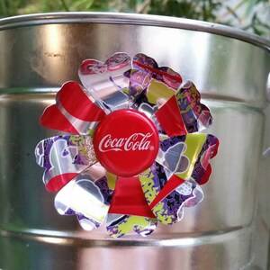 Puede incluir: Un cubo de metal plateado con una flor roja, plateada y morada hecha de latas de aluminio recicladas. El centro de la flor es una tapa de botella de Coca-Cola roja.