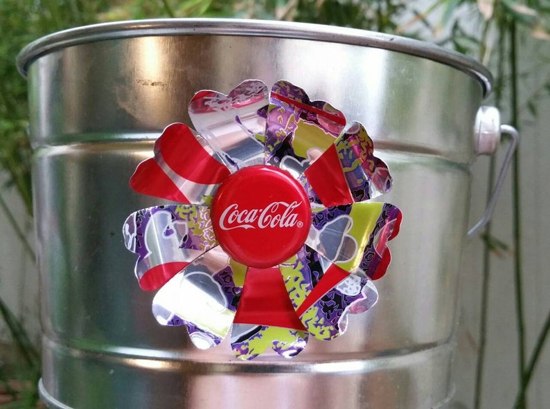 Puede incluir: Un cubo de metal plateado con una flor roja, plateada y morada hecha de latas de aluminio recicladas. El centro de la flor es una tapa de botella de Coca-Cola roja.