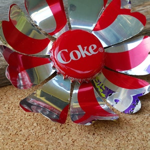 Puede incluir: Una flor roja y plateada hecha de leng&uuml;etas de latas de refresco de aluminio. El centro de la flor es una tapa de botella de Coca-Cola roja con la palabra "Coke" impresa en blanco.