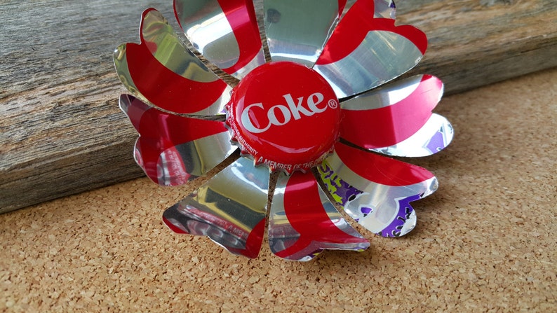 Puede incluir: Una flor roja y plateada hecha de leng&uuml;etas de latas de refresco de aluminio. El centro de la flor es una tapa de botella de Coca-Cola roja con la palabra "Coke" impresa en blanco.