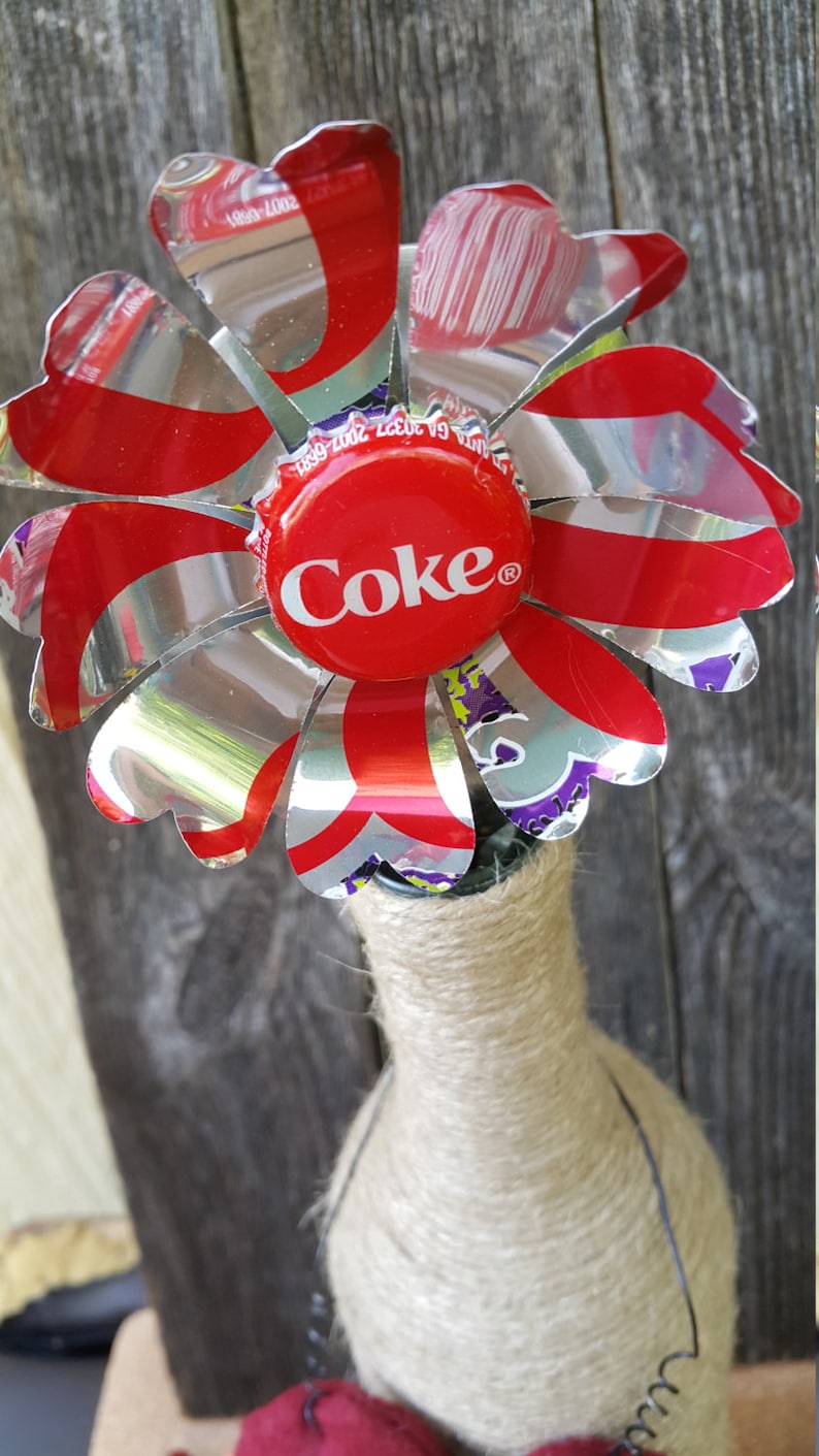 Puede incluir: Una flor roja y plateada hecha de latas de refresco recicladas con una tapa de botella de Coca-Cola en el centro. La flor est&aacute; unida a una botella envuelta en cuerda.