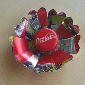 Puede incluir: Una flor roja, blanca y plateada hecha de latas de aluminio con una tapa de botella de Coca-Cola en el centro.