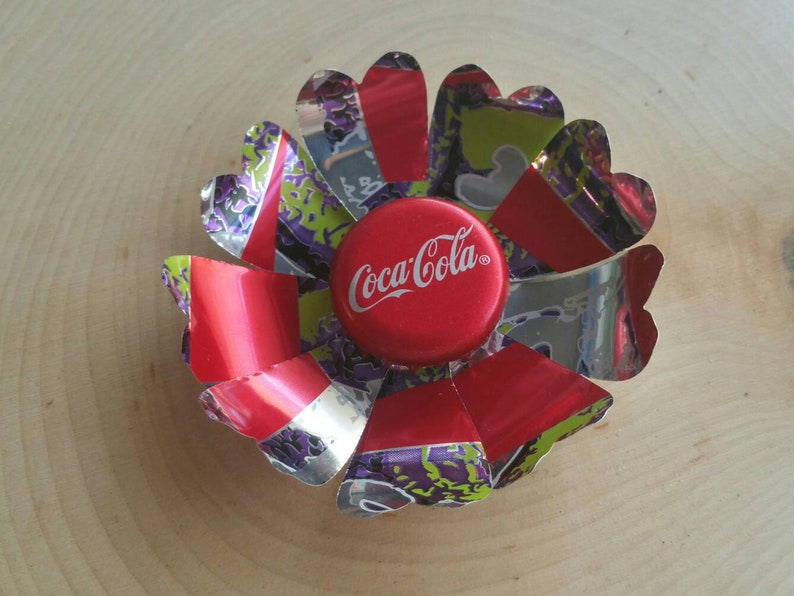 Puede incluir: Una flor roja, blanca y plateada hecha de latas de aluminio con una tapa de botella de Coca-Cola en el centro.
