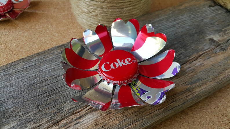 Puede incluir: Una flor roja y plateada hecha de una lata de refresco con una tapa de botella de Coca-Cola roja en el centro.