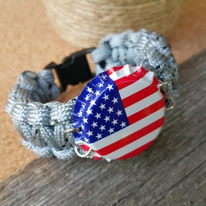 K&ouml;nnte beinhalten: Ein graues Paracord-Armband mit einer schwarzen Schnalle und einem Flaschenverschluss mit der amerikanischen Flagge.