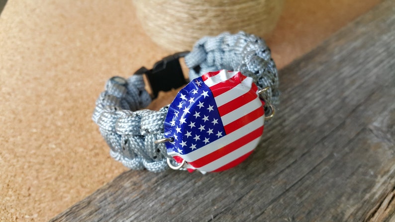 K&ouml;nnte beinhalten: Ein graues Paracord-Armband mit einer schwarzen Schnalle und einem Flaschenverschluss mit der amerikanischen Flagge.
