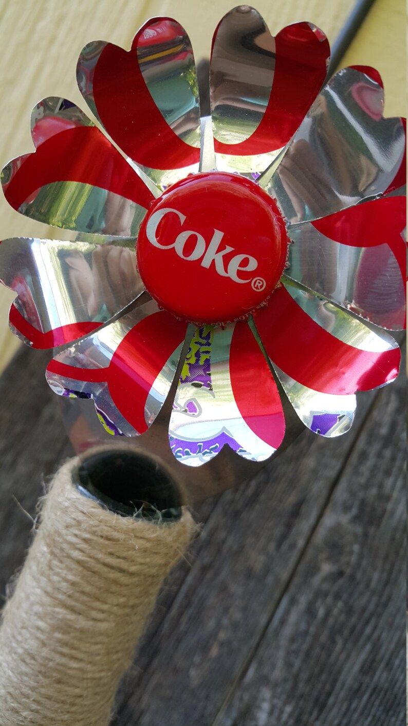 Puede incluir: Una flor roja y plateada hecha de latas de aluminio recicladas con una tapa de botella de Coca-Cola roja en el centro.