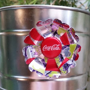Puede incluir: Un cubo de metal plateado con una flor colorida hecha de latas de aluminio recicladas. La flor tiene un tap&oacute;n de botella de Coca-Cola rojo en el centro.